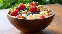 Una imagen vibrante y atractiva de un açai bowl servido en un tazón rústico de madera, decorado con frutas frescas como fresas, plátano, arándanos y granola. El fondo muestra un ambiente natural con hojas verdes, evocando la selva amazónica. Los colores deben ser intensos y llamativos, resaltando el morado profundo del açai. La imagen debe transmitir salud, frescura y energía, ideal para una publicación sobre los beneficios de este súper alimento.