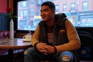 Un joven migrante venezolano, Romeo 2.0, está sentado en un pequeño café en el Bronx, Nueva York, en un ambiente acogedor con luces tenues y decoración romántica por San Valentín. Tiene unos 25 años, piel trigueña, cabello negro corto y viste una chaqueta casual con jeans. En la mesa hay una taza de café humeante y un teléfono con una conversación de WhatsApp abierta. Frente a él, un periodista con libreta en mano lo entrevista. Romeo sonríe con nostalgia mientras habla, mirando hacia la ventana donde se ven edificios de ladrillo típicos del Bronx y algunas luces de neón reflejadas en el vidrio. El ambiente es invernal, con personas caminando afuera con abrigos. Pequeños detalles como una rosa en la mesa y una bandera venezolana en su mochila agregan profundidad a la historia. Estilo realista con tonos cálidos y detalles cinematográficos.